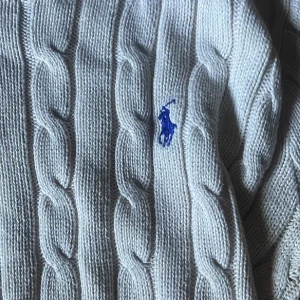 Vit kabelstickad tröja från Ralph Lauren - Vit kabelstickad tröja från Ralph Lauren med klassisk blå broderad logga på bröstet. Tröjan har rund halsringning, långa ärmar och ribbade muddar. Perfekt för dig som gillar en clean och tidlös stil.