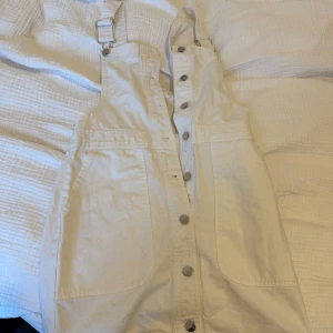 Vit kort klänning från mango Casual - Supersöt vit kort klänning från MNG Casual i jeansstil med knappar framtill och justerbara axelband. Klänningen har stora fickor.