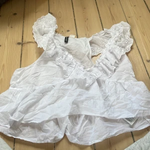 Vit volangtopp från H&M Divided - Supersöt vit topp från H&M Divided med volanger och broderade spetsdetaljer vid axlarna. Toppen är ärmlös och har en luftig, flowy passform som känns perfekt för varma dagar. Tillverkad i bomull för en skön känsla.