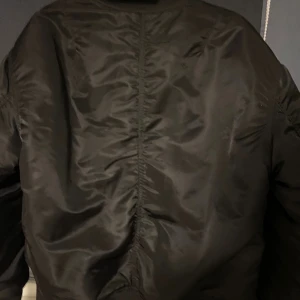 Svart bomberjacka från H&M - Snygg svart bomberjacka från H&M med klassisk siluett, ribbade muddar och silverfärgad dragkedja. Jackan har två snedställda fickor framtill och en ärmficka med dragkedja. Tillverkad i glansig polyester som ger en cool look.