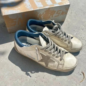 Golden Goose vita sneakers med blå detalj - Sneakers från Golden Goose. Super-Star från Golden Goose förenar en vintageinspirerad design med unika patinaeffekter och den ikoniska stjärnan på utsidan. Skorna är väl använda men har mer kvar att ge. Perfekta sommarskor för en cool avslappnad look. Kan även gå ner i pris vid snabb affär. 