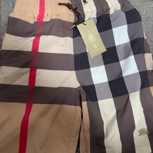 Burberry shorts - Säljer ett par klassiska Burberry shorts med stort rutigt mönster i beige, brunt, svart, vitt och en röd rand. Shortsen har snörning i midjan och är perfekta för sommaren. Ikonisk design och riktigt snygga detaljer.