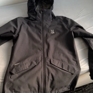 Haglöfs jacka  - Svart vindjacka från Haglöfs med huva, hel dragkedja och två stora fickor framtill. Jackan har meshfoder på insidan och elastiska muddar vid ärmsluten. Perfekt för blåsiga dagar och outdoor-äventyr.