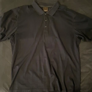 Pikétröja från Oscar Jacobson - Väldigt mörk blå (nästan svart) klassisk pikétröja från Oscar Jacobson med krage och knappar framtill. Tillverkad i mjuk bomull och har diskret broderad logga nedtill. Perfekt till jeans eller chinos för en clean look.