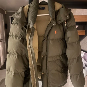 Olivgrön pufferjacka Polo Ralph Lauren - Olivgrön pufferjacka från Polo Ralph Lauren med orange broderad logga på bröstet. Jackan har huva, dragkedja och tryckknappar framtill samt två stora fickor. Insidan är beige och jackan är vadderad för extra värme. Perfekt för kalla dagar.
