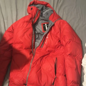 Röd dunjacka från Sail Racing S - Riktigt fet röd dunjacka från Sail Racing i storlek S. Jackan har en clean design med grått innerfoder, dragkedja framtill och logga på bröstet och ryggen. Perfekt för kalla dagar med sin fluffiga och varma känsla. Snygg och sportig look.