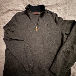 Mörkgrå halvzip-tröja från Morris - Mörkgrå stickad tröja från Morris i 100% merinoull med halv dragkedja och hög krage. Klassisk och stilren design med diskret broderad logga på bröstet. Perfekt för lager-på-lager och snygg till jeans eller chinos. Storlek S