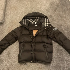 Svart pufferjacka från Burberry - Svart pufferjacka från Burberry med avtagbara ärmar så du kan bära den som väst. Jackan har huva med klassiskt rutigt Burberry-foder i grått och vitt, dragkedja och knappar framtill samt två fickor med dragkedja.