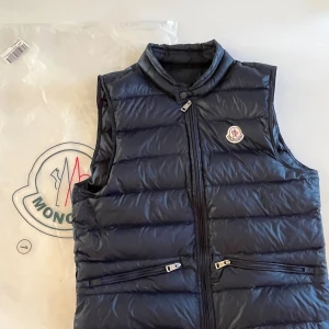Mörkblå dunväst från Moncler - Snygg mörkblå dunväst från Moncler med hög krage och dragkedja framtill. Västen har två snedställda fickor med dragkedja och klassisk Moncler-logga på bröstet. Perfekt för lager-på-lager och streetstyle. Originalpåse medföljer.