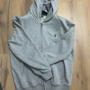 Grå hoodie från Polo Ralph Lauren - Snygg grå hoodie från Polo Ralph Lauren med dragkedja och klassisk broderad logga på bröstet. Tröjan har huva, långa ärmar och ribbade muddar. Perfekt för chill dagar och enkel att matcha med jeans eller joggers.