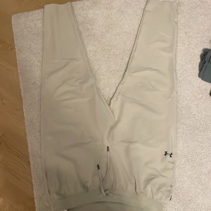 Beige mjukisbyxor från Under Armour - Beiga mjukisbyxor från Under Armour med dragsko i midjan och resår vid benslut. Byxorna har en enkel design med diskret logga på ena benet och är tillverkade i ett mjukt syntetmaterial som passar perfekt för chill eller träning.