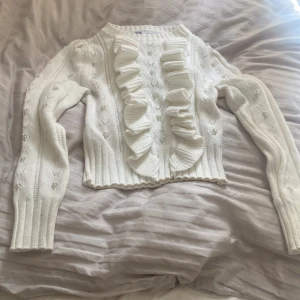 Vit stickad tröja med volang från Zara - Supersöt vit stickad tröja från Zara med volangdetaljer framtill och pärlor på ärmarna. Tröjan har hjärtmönster, ribbade muddar och lång ärm. Använd endast en gång 
