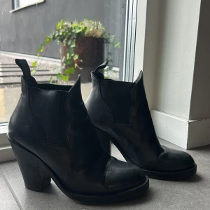 Svarta boots med klack från Acne - Säljer ett par svarta boots från Acne med markerad klack och rund tå. Skorna är i skinn och har snygga detaljer längs skaftet som ger en cool western-vibe. Perfekta för dig som vill ha en stilren men edgy look.