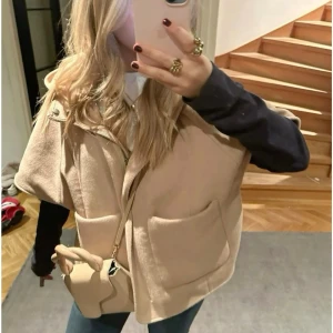 Beige cape/poncho med huva från Zara - Stilren beige capeväst från Zara med huva och två stora fickor framtill, dragkedja och tryckknapp vid halsen. Perfekt för lager-på-lager och nu till hösten. Från barnavdelningen och är i Storlek 140 men passar xs/s 😊