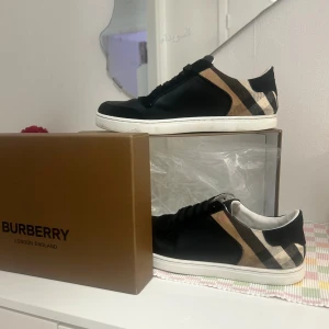Burberry sneakers med klassiskt rutmönster - Snygga svarta sneakers från Burberry med klassiskt beige och svart rutmönster på sidorna. Skorna har vita sulor, svarta skosnören och detaljer i både skinn och textil. Perfekta för dig som vill ha en stilren och lyxig look med ikoniska Burberry-detaljer.