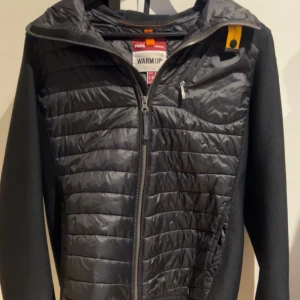 Svart quiltad jacka Parajumpers Y-M/S - Snygg svart jacka från Parajumpers i storlek Y-M. Jackan har quiltade partier framtill och baktill, huva och dragkedja. Ärmarna är i ett stickat material och jackan har en gul detalj vid kragen. Perfekt för dig som vill ha en sportig och stilren look. Pris kan diskuteras vid en snabb affär 