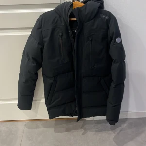 Svart parkajacka från Sail Racing L - Parkajacka från Sail Racing i svart, modell Race T8 Parka. Jackan har huva, två bröstfickor med dragkedja och vadderad design. Snygg logga på bröstet och ärmen. Perfekt för kalla dagar och riktigt stilren look. helt ny med prislappen kvar, aldrig använd 