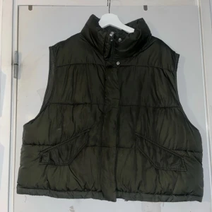 Dunväst - Säljer en mörkgrön dunväst med hög krage och dragkedja framtill. Västen har två snedställda fickor med lock och quiltad design. Perfekt för lager-på-lager och ger en cool, avslappnad look. Passar dig som gillar streetstyle och vill hålla dig varm.