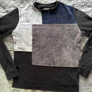 Blockmönstrad tröja från Le Vichy - Snygg långärmad tröja från Le Vichy med blockmönster i svart, grått, vitt och mörkblått. Tröjan har en cool patch på ena ärmen och är gjord i mjuk bomull och sammet. Perfekt för dig som gillar unika detaljer och streetstyle.