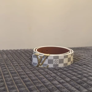 Vit-guld bälte  - Stilrent bälte med rutigt mönster i ljusgrått och vitt. Spännet är i blank guldfärg och har en elegant, modern design. Insidan är i ljusbrunt läder som ger en fin kontrast mot den ljusa utsidan. Ett tidlöst accessoarval som passar både till vardags och festligare tillfällen.