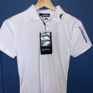 Vit pikétröja från J.Lindeberg - Snygg vit pikétröja från J.Lindeberg med klassisk krage och korta ärmar. Tröjan har svart logga broderad på kragen och ärmen samt diskreta knappar framtill. Perfekt för golf eller en clean look. Helt ny helt oanvänd.