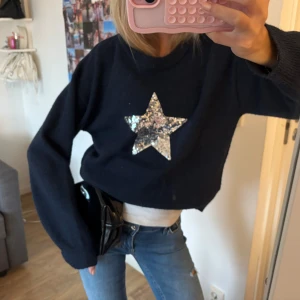 Mörkblå stickad tröja med paljettstjärna - Mysig mörkblå stickad tröja med en stor silvrig paljettstjärna på bröstet. Tröjan har en rund halsringning och långa ärmar. Perfekt för dig som vill sticka ut lite extra med en glittrig detalj.