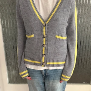 Grå kofta med gula ränder från Zara - Grå stickad kofta från Zara med gula och vita ränder längs kanter, ärmslut och fickor. Djup v-ringning, knappar framtill och två små fickor. Perfekt att slänga över en t-shirt för en chill look. Så cool och unik, knappt använd. 