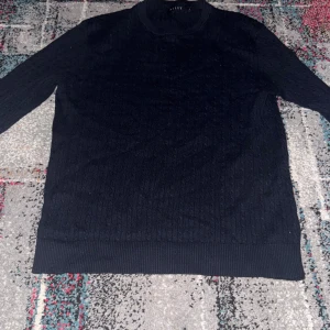 Mörkblå kabelstickad tröja från Riley - Kabelstickad mörkblå tröja från Riley med rund halsringning och ribbade muddar. Tröjan har lång ärm och klassisk kabelmönster, perfekt för lager-på-lager under kyliga dagar. Enkel och stilren design som funkar till det mesta.