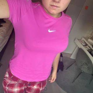 Rosa Nike t-shirt i sportig stil - Säljer en rosa t-shirt från Nike med klassisk vit logga på bröstet. T-shirten har korta ärmar och rund halsringning. Perfekt för träning eller en aktiv vardag. Materialet känns lätt och andas bra.