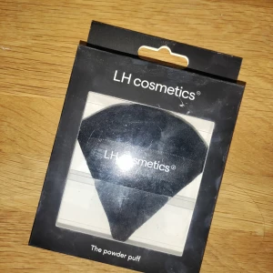 Svart powder puff från LH cosmetics - En mjuk och svart powder puff från LH cosmetics, perfekt för att applicera och bättra på puder. Puffen har en trekantig form med ett band för grepp där LH cosmetics-loggan syns. Materialet är lent och designen gör det enkelt att nå alla ansiktskonturer. Aldrig använd 