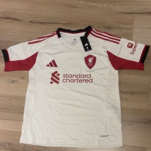 Liverpool FC vit och röd matchtröja Adidas - Liverpool FC:s officiella bortatröja från Adidas i vitt med röda detaljer och klubbmärke på bröstet. Tröjan har korta ärmar, svarta och röda ränder på axlarna samt tryckta sponsorer. Tillverkad i lätt och ventilerande polyester.