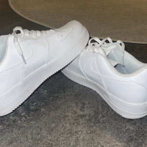 Nike Air Force 1 helvita sneakers - Klassiska Nike Air Force 1 sneakers i helvitt skinn med vit sula och snörning. Snygg och clean design med perforerad tå och ikoniska swoosh-loggan på sidorna. Perfekta för dig som gillar stilrena och tidlösa sneakers.