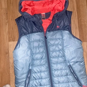 Blå dunväst med huva från Quechua - Snygg dunväst från Quechua i ljusblå och mörkblå nyans med orange foder. Västen har huva, dragkedja framtill och två fickor med dragkedja. Perfekt för lager-på-lager och outdoor-aktiviteter.