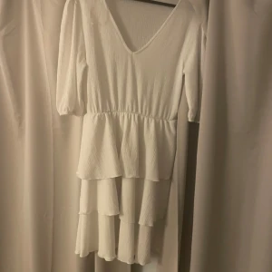 Vit volangklänning med v-ringning - Supersöt vit klänning från Bik Bok med v-ringad hals och trekvartsärmar. Klänningen har flera lager volanger och är tillverkad i ett lätt, strukturerat tyg som ger en skön känsla. Perfekt för dig som gillar romantisk och feminin stil.