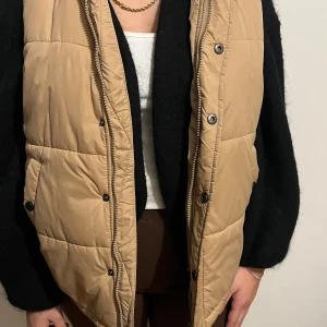 Beige/ ljusbrun väst  - En beige/ ljusbrun quiltad väst med knappar framtill. Den har två fickor vid sidorna med knappar på. Lätt väst som inte är tung. Rak i passformen och lite oversized. Västen har även en luva till. 