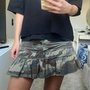 Camo minikjol med volang och knappar - Säljer en kort camouflagekjol med volangkant och flera knappar framtill. Kjolen har dragkedjor på sidorna och coola detaljer som ger en edgy vibe. Perfekt för dig som vill sticka ut med en streetstyle-look.