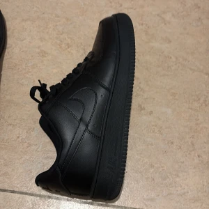 Svarta Nike Air Force 1 sneakers - Säljer ett par helsvarta Nike Air Force 1 sneakers i skinn med klassisk siluett och perforerad tå. Skorna har snörning, metallbricka med AF1-logga och diskret Nike Swoosh på sidorna. Perfekta för dig som gillar en clean och tidlös look.