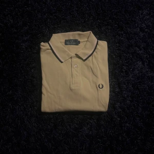 Pikétröja från Fred Perry - Snygg beige pikétröja från Fred Perry med klassisk krage och två knappar. Tröjan har svart och vit detalj på kragen samt den ikoniska Fred Perry-loggan broderad på bröstet. Tillverkad i 100% bomull för en skön och luftig känsla.