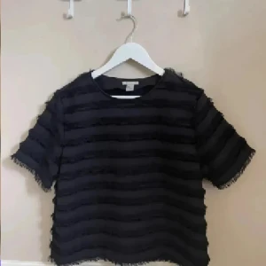 Svart randig blus med fransiga detaljer - Säljer en svart blus från H&M med horisontella ränder och fransiga kanter. Blusen har rund halsringning och korta ärmar. Materialet känns lätt och luftigt, perfekt för dig som gillar en cool och unik stil. Passar dig som vill sticka ut lite extra.