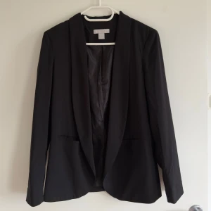 Fin svart kavaj från H&M, strl 34 - Snygg svart kavaj från H&M i storlek 34. Kavajen har klassisk design med lång ärm, två fickor framtill och mjukt foder. Materialet är 92% polyester och 8% elastan, fodret är 100% polyester. Perfekt för dig som gillar stilrena och enkla plagg.
