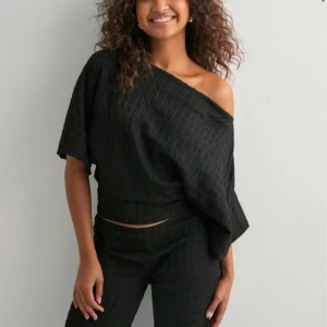 Svart offshoulder topp - Säljer en svart offshoulder topp med lös passform och kort ärm. Toppen har en snygg struktur i tyget och sitter skönt över ena axeln. (Har byxorna om det skulle vara intressant så blir det ett set). Storlek xs. Från Nelly. 