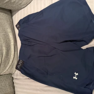 Blå träningsshorts från Under Armour - Säljer ett par marinblå träningsshorts från Under Armour med elastisk midja och logga på benet. Perfekta för gymmet eller löprundan, lätta och bekväma med sportig look.