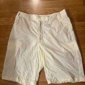 Vita cargo shorts med fickor - Säljer ett par vita cargo shorts med flera praktiska fickor och bälteshällor. De har knappstängning framtill och en metallögla för extra detalj. Perfekta för sommardagar och chill stil. Materialet känns lätt och luftigt.