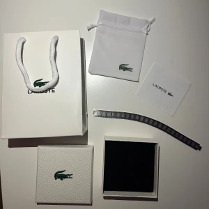 Silverfärgat armband från Lacoste - Stilrent armband från Lacoste i silverfärgad metall med länkdesign och ingraverade Lacoste-loggor på varje länk. Snyggt krokodilmotiv på spännet. Levereras med original ask, påse och instruktioner. Perfekt accessoar för dig som gillar klassisk och sportig stil.