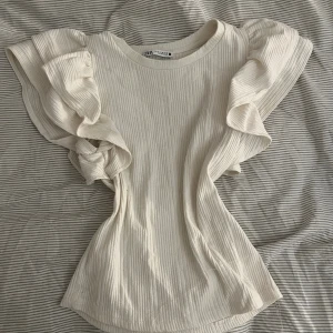 Ribbad volangblus från Zara - Säljer en ribbad, ljusbeige blus från Zara med volangärmar. Toppen har rund halsringning och är tillverkad i ett mjukt bomullsmaterial. Perfekt för dig som gillar en feminin och trendig look med extra volym på axlarna.