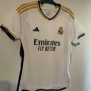 Real Madrid hemmatröja Adidas L - Säljer en  Real Madrid hemmatröja från Adidas i storlek L. Tröjan är vit med guld- och marinblå detaljer, broderat klubbmärke och svarta Adidas-loggor. perfekt för match eller träning.