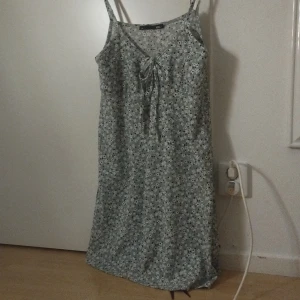 Blommig sommarklänning från 157 - Ljusgrön blommig klänning från 157 i storlek L. Klänningen har smala axelband, v-ringning med knyt framtill och ett sött småblommigt mönster i vitt och svart. Perfekt för varma dagar och har en avslappnad passform.