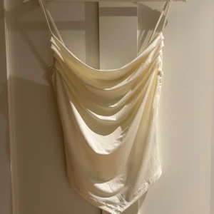 Krämvit linne från Bik Bok - Säljer en krämvit linne från Bik Bok i storlek S. Toppen har tunna justerbara axelband och en snygg drapering framtill som ger en cool och trendig look. Materialet är glansigt och känns lyxigt mot huden, perfekt för att lyfta din outfit.