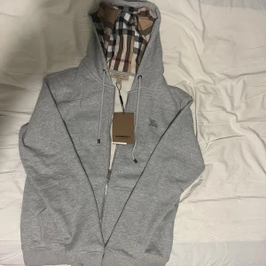 Grå hoodie från Burberry XL - Snygg grå hoodie från Burberry i storlek XL med klassiskt rutigt foder i huvan och insidan. Tröjan har dragkedja, snörning i huvan och broderad logga på bröstet. Perfekt för dig som vill ha en stilren och lyxig look. Det är inte en riktig XL det är en L