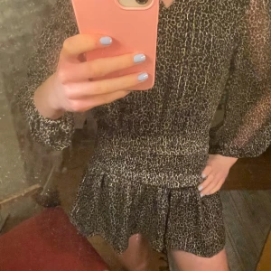 Kort leopardklänning med guldtrådar - Snygg kort klänning med leopardmönster och diskreta guldtrådar. Klänningen har v-ringning, långa transparenta ärmar och smockad midja som ger en markerad siluett. Kjolen har volangkant.